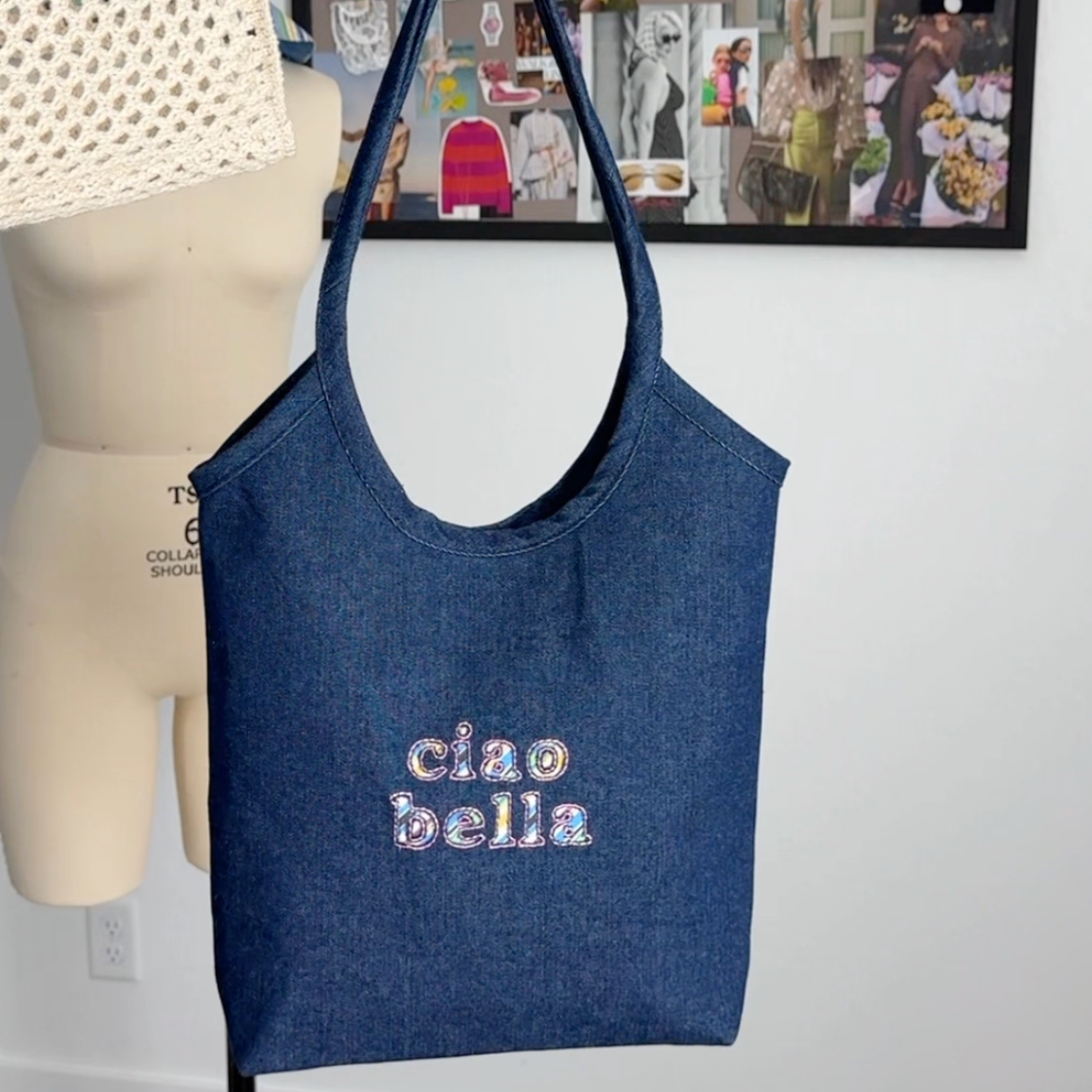 A navy blue denim tote bag with 'ciao bella' embroidered on the front, held by a hand in front of a mannequin torso and a framed collage on a white wall.