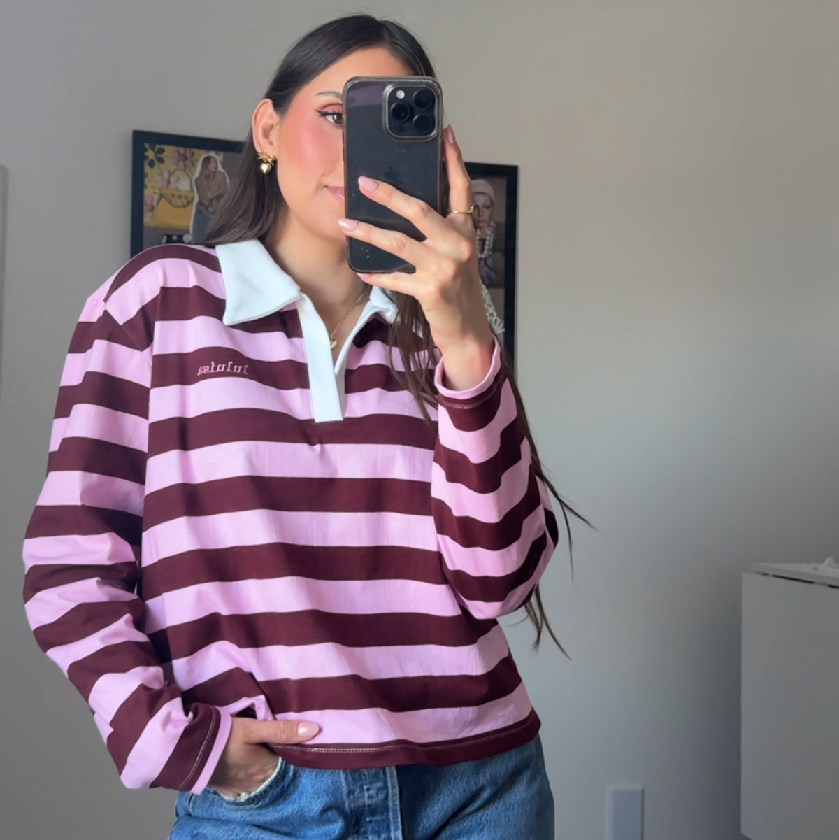 A woman takes a mirror selfie wearing a pink and burgundy striped long-sleeve polo shirt with a white collar, paired with blue jeans, in an indoor room setting.