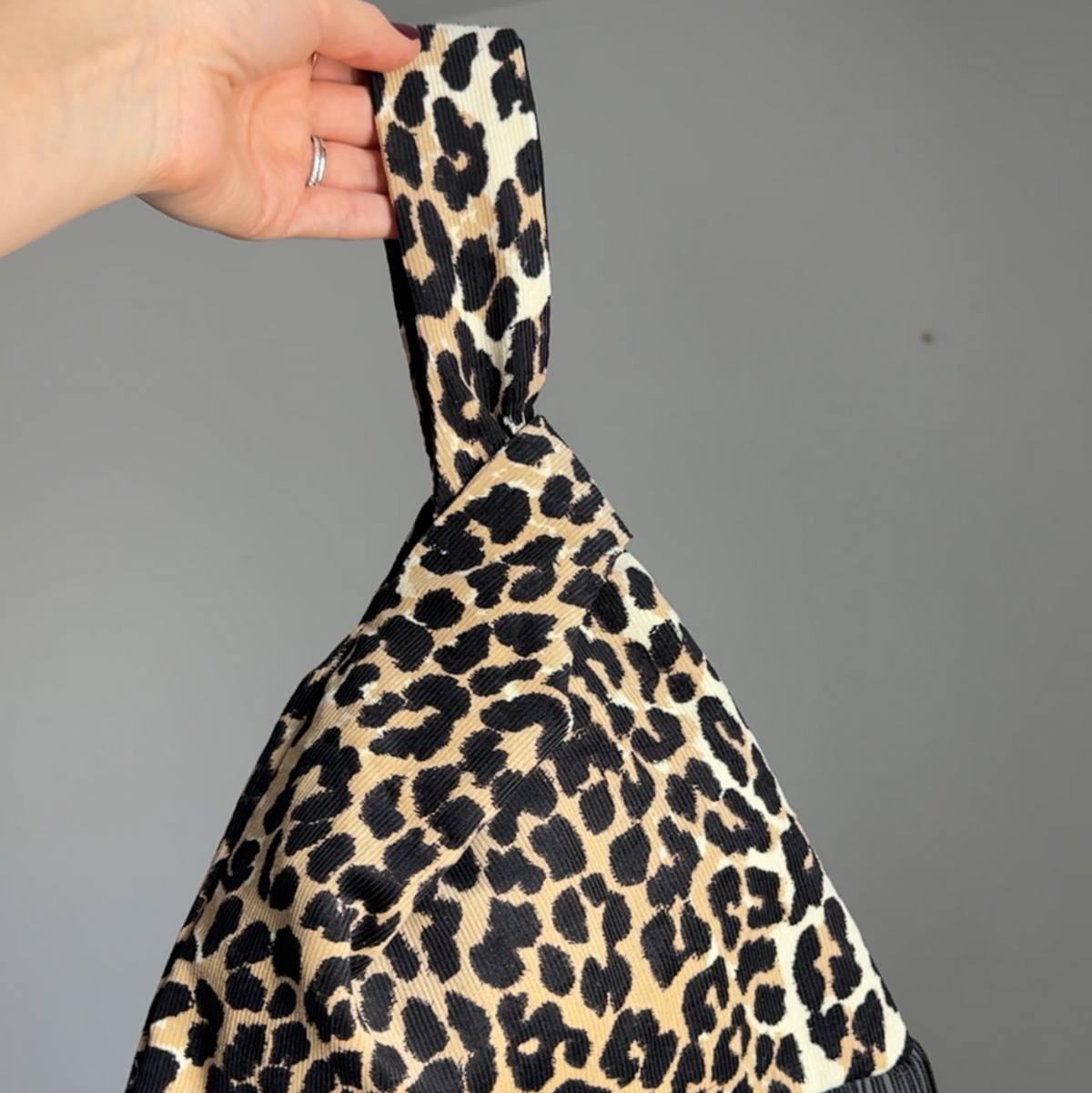 A leopard print japanese knot handbag with black fringe, held up by a hand against a plain gray wall.