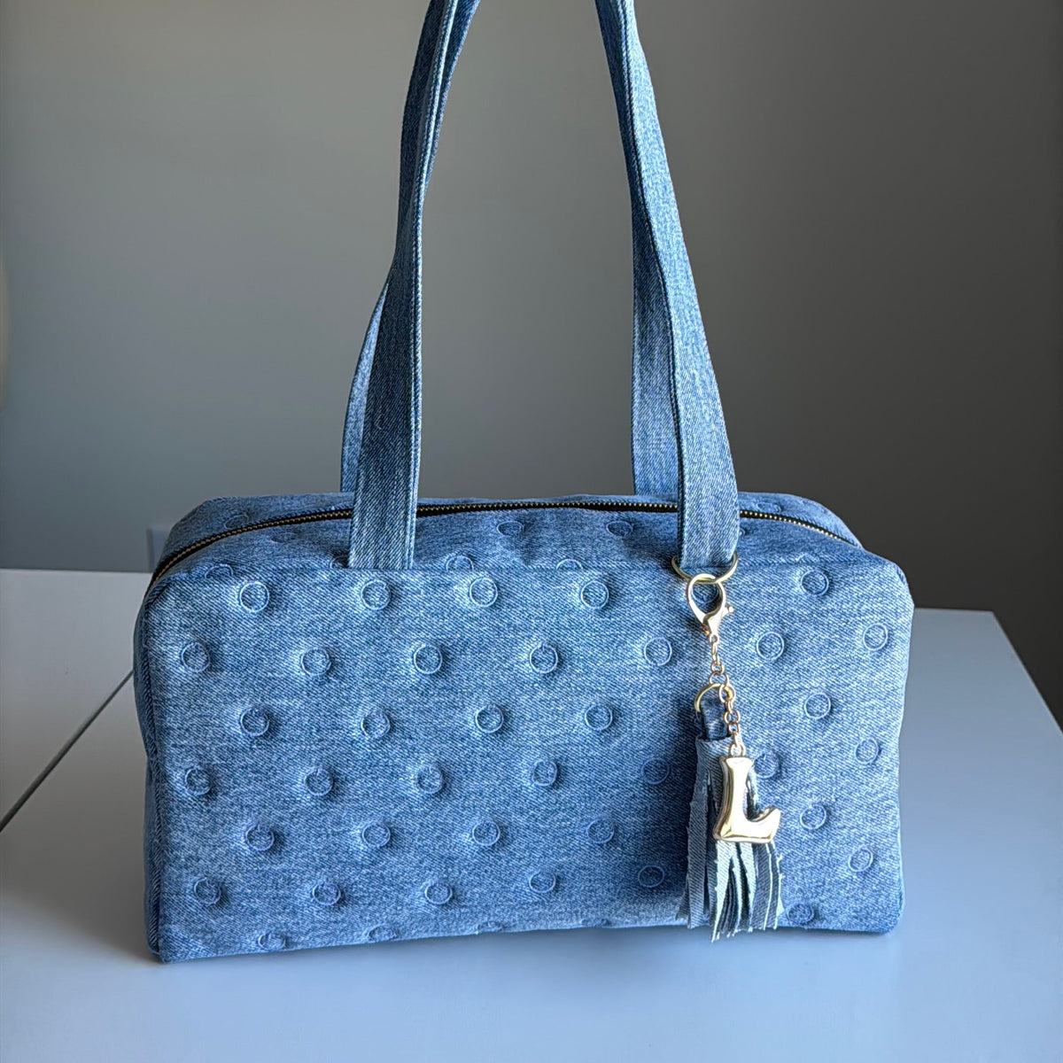 A blue ostrich-textured leather handbag with dual top handles and a silver-toned tassel charm, displayed on a light gray surface against a neutral gray background.