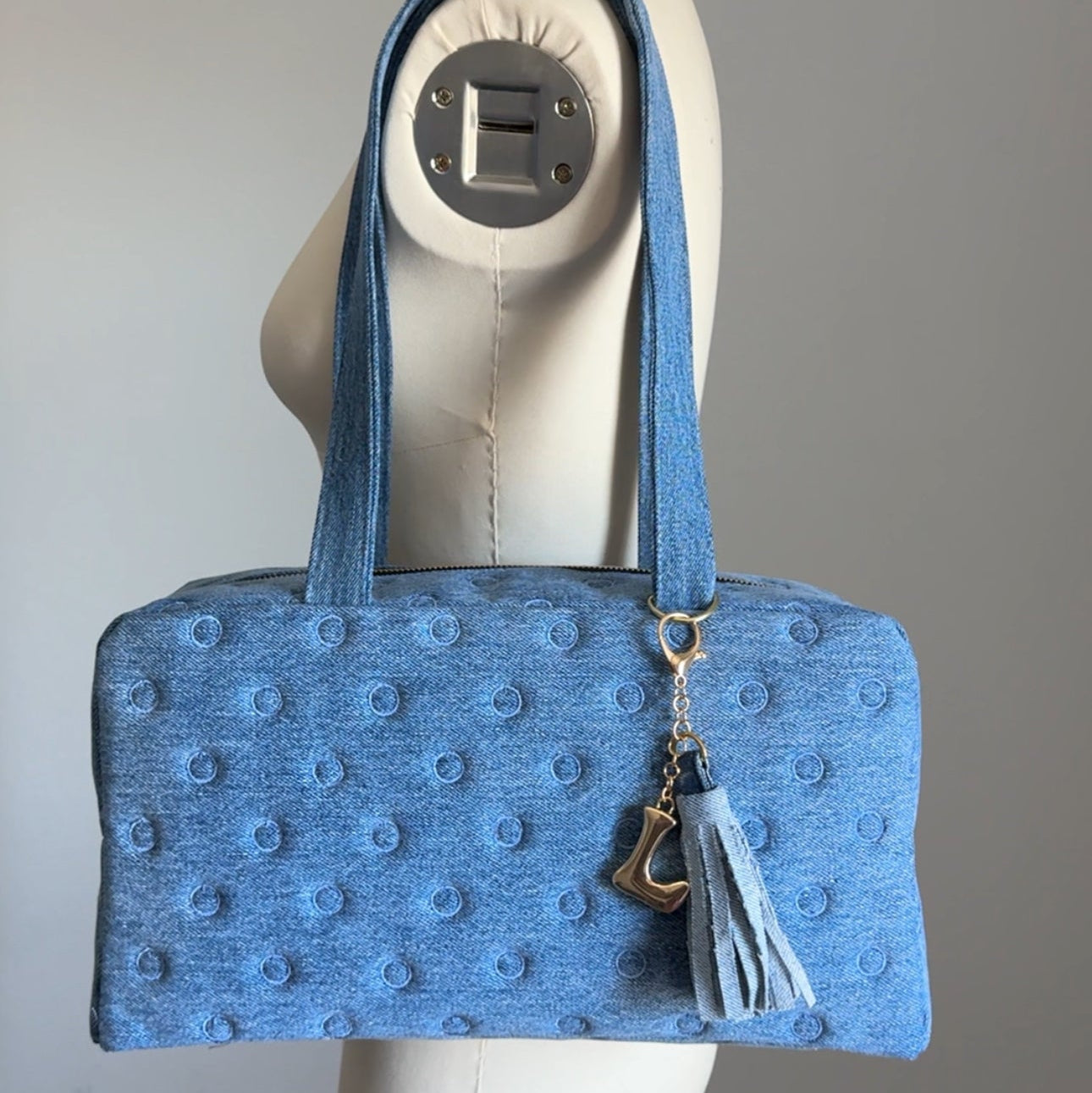 A light blue textured bowler-style bag with matching shoulder straps, with polka dots embroidered, displayed on a cream dress form mannequin. The bag features a textured, dotted fabric pattern, a gold-tone keychain ring, and a light blue tassel charm.