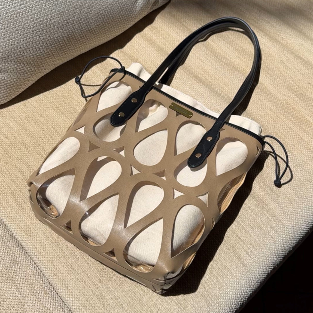 A structured tote bag with a beige geometric cutout pattern over white fabric lining, featuring black leather handles and drawstring details, resting on a textured beige fabric surface.