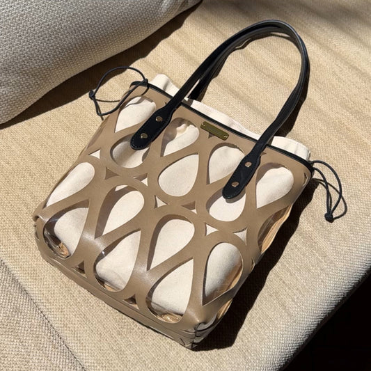 A structured tote bag with a beige geometric cutout pattern over white fabric lining, featuring black leather handles and drawstring details, resting on a textured beige fabric surface.