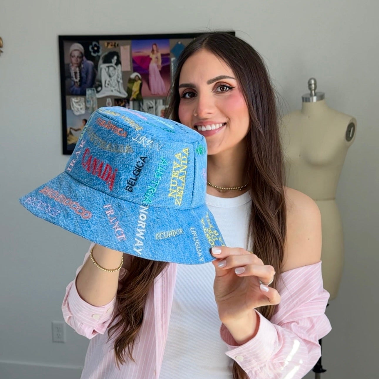 Woman holding a blue hat with colorful text in a room with a mannequin and framed pictures.