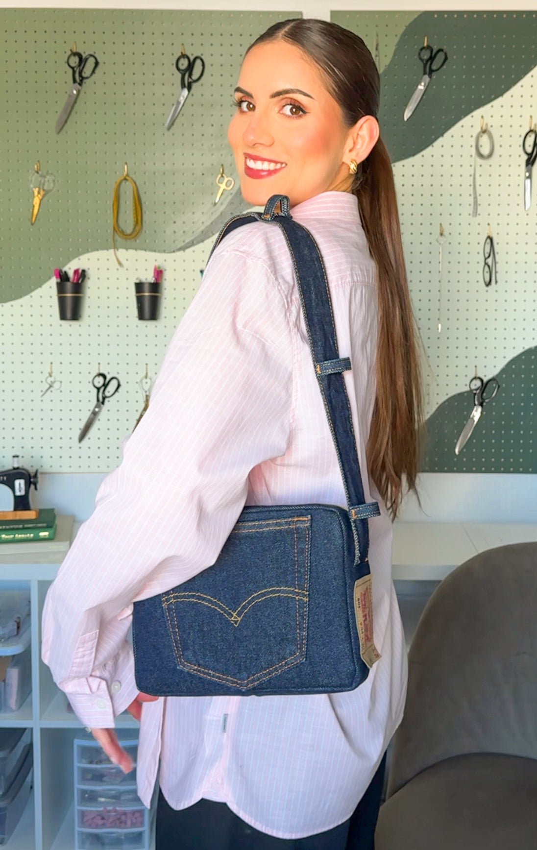 Woman wearing a denim crossbody bag in a workshop setting with tools on the wall. Shoulder bag levis jeans upcycle pattern, bolsa de hombro rectangular con jeans y bolsillos de levis usando la pretina, waist ban shoulder bag levis upcycled denim jeans