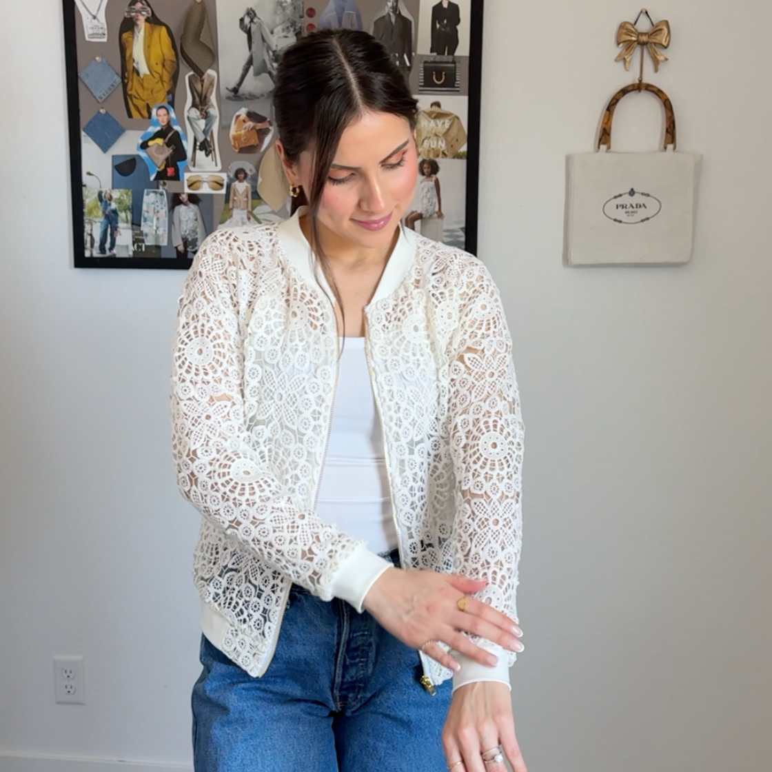 Woman wearing a white lace cardigan over a white shirt and blue jeans, standing in a room with a wall collage and handbag.