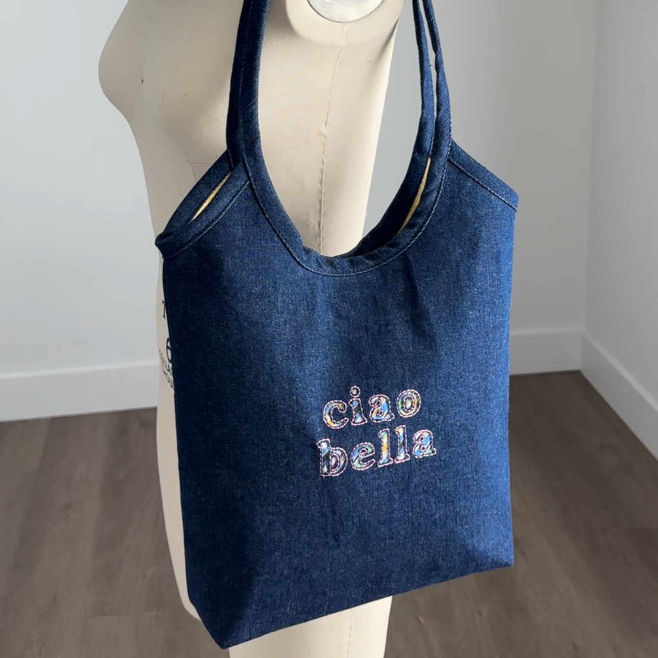 A blue denim tote bag with 'ciao bella' embroidered on the front, displayed on a cream-colored dress form mannequin in an indoor room setting.
