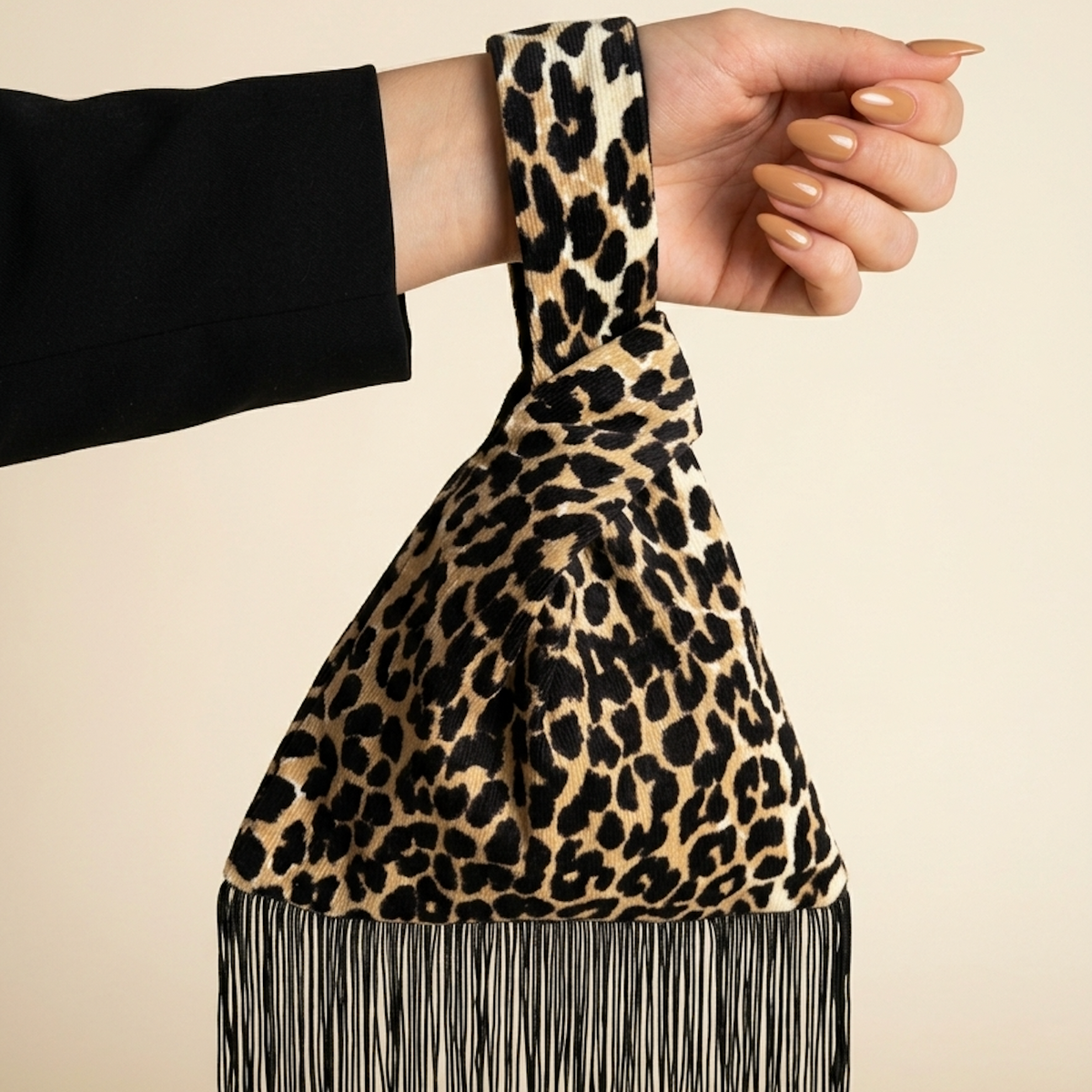 A leopard print shoulder bag with a wide strap and fringe hem, held by a person's hand against a solid beige background.